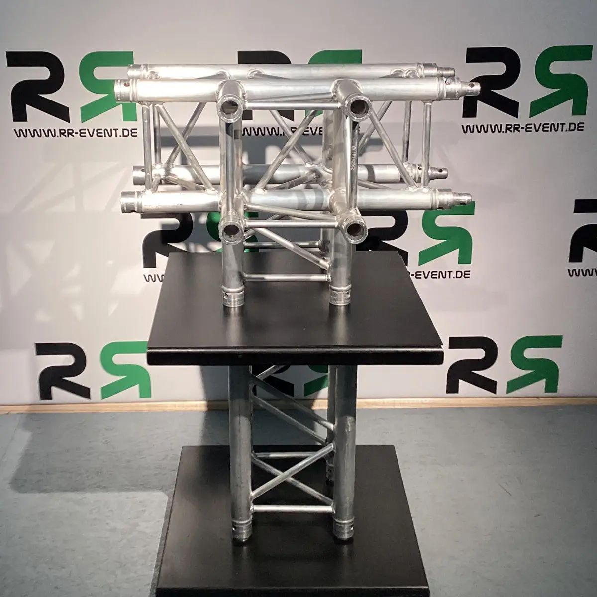 Die abgebildete Vierpunkttraverse aus Oldenburg, die auf einem schwarzen Podest vor einer weißen Wand mit RR-EVENT.DE-Logos präsentiert wird, eignet sich ideal zum Verleih für Veranstaltungen und sorgt für eine stabile sowie flexible Bühnenkonstruktion.
